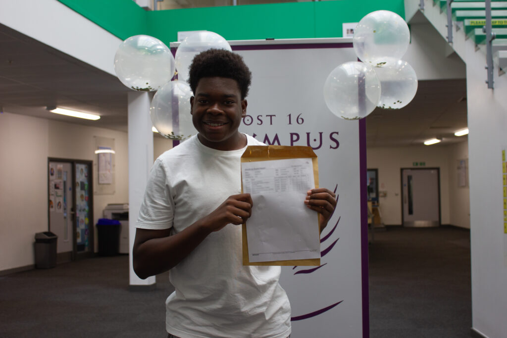 A male Post-16 student is pictured smiling for the camera, whilst holding his A-Level results in his hand.