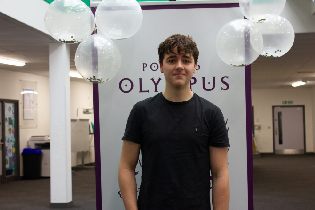A male Post-16 student is pictured smiling for the camera, whilst holding his A-Level results in his hand.
