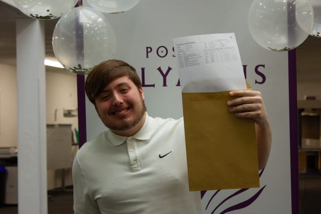 A male Post-16 student is pictured smiling for the camera, whilst holding his A-Level results in his hand.