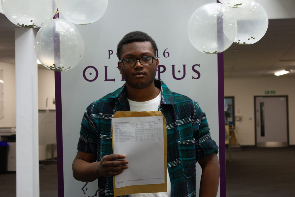 A male Post-16 student is pictured smiling for the camera, whilst holding his A-Level results in his hand.