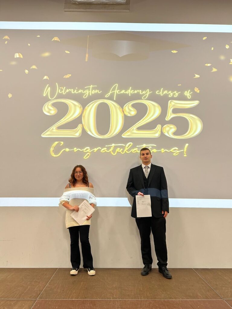 Two students standing on the stage in front of the same "Wilmington Academy class of 2025 Congratulations!" display, proudly holding their certificates. They are likely posing for individual or small group portraits during the ceremony.