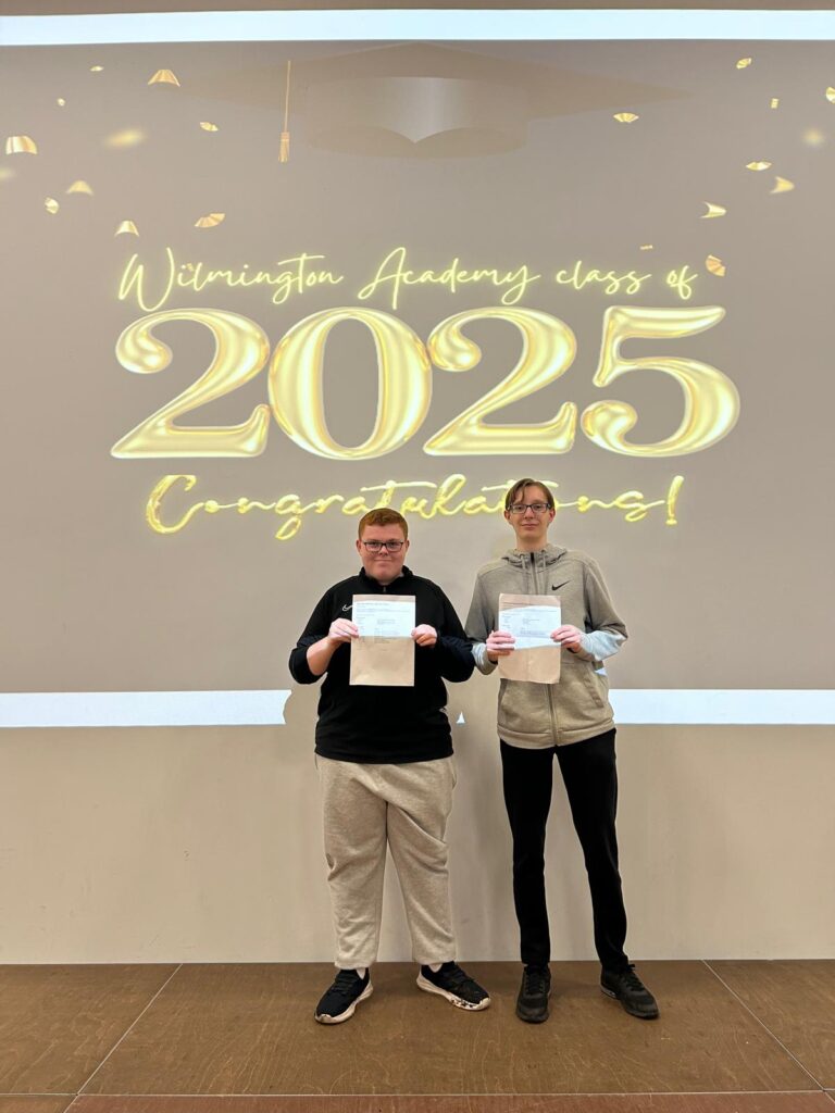 Two students standing on the stage in front of the same "Wilmington Academy class of 2025 Congratulations!" display, proudly holding their certificates. They are likely posing for individual or small group portraits during the ceremony.