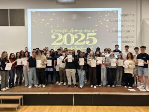 A shot of students on the stage, all holding their certificates and smiling. The "Leigh Academy Wilmington class of 2025 Congratulations!" message is prominent on the screen behind them. On the right side of the wall, there are words like "inquirers," "knowledgeable," "thinkers," "communicators," "principled," and "open-minded," which might be related to the academy's values.