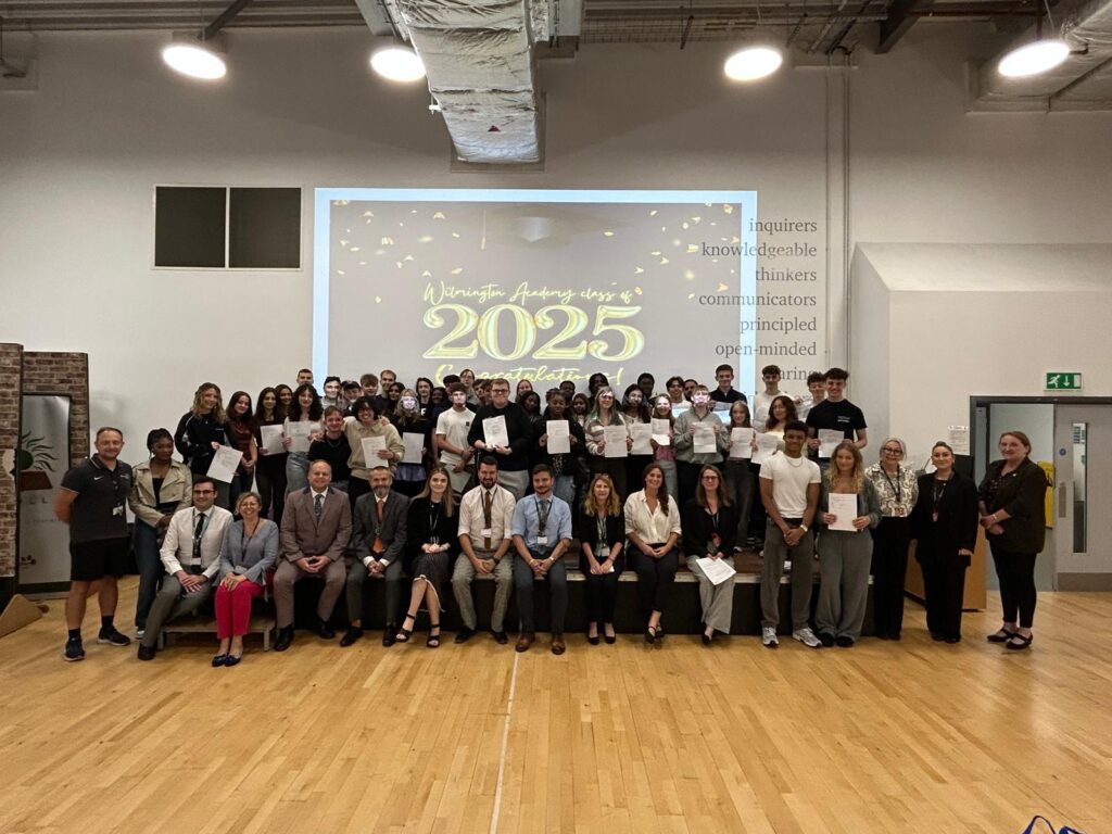 A large group of students, likely the graduating class of 2025 from Wilmington Academy, are gathered on a stage with some adults (presumably faculty or staff) seated in the front row. Most of the students are holding certificates or papers. A large screen behind them displays "Wilmington Academy class of 2025 Congratulations!" with a graduation cap and falling confetti animation.