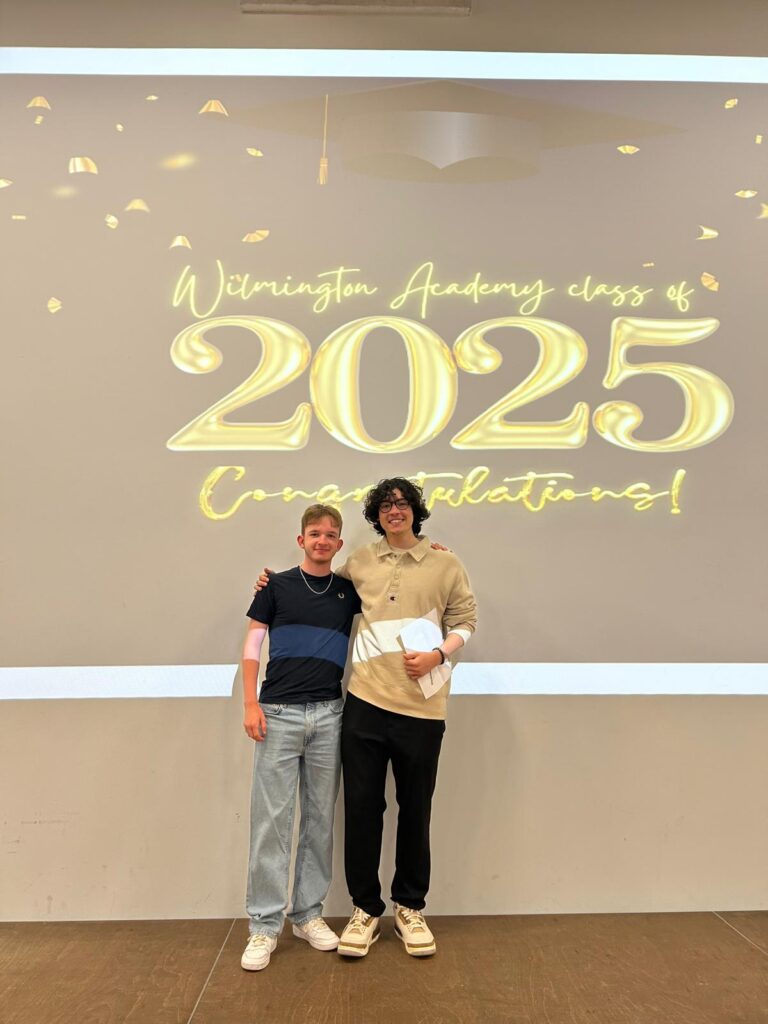 Two students standing on the stage in front of the same "Wilmington Academy class of 2025 Congratulations!" display, proudly holding their certificates. They are likely posing for individual or small group portraits during the ceremony.