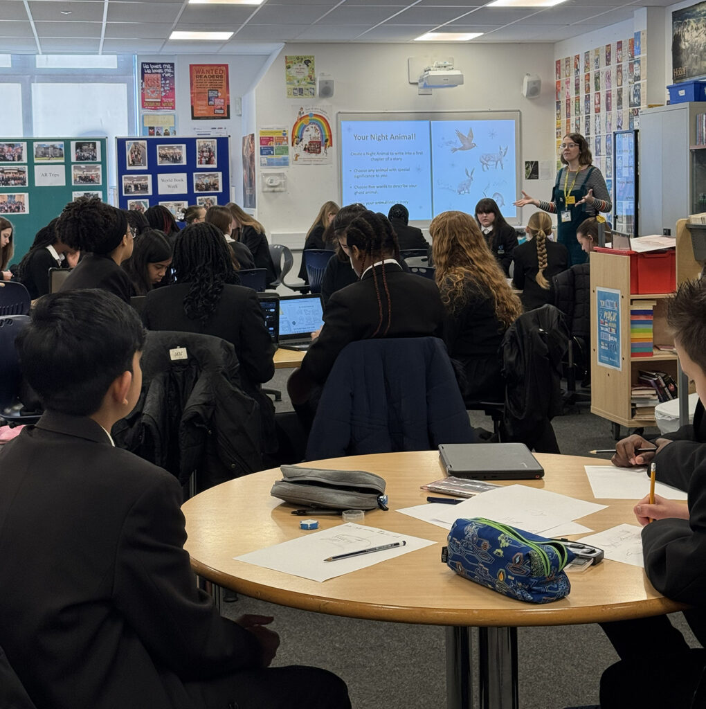 A speaker presents a slide titled "A little about me," featuring author Sarah Ann Juckes and a list of her books, to a classroom of students.