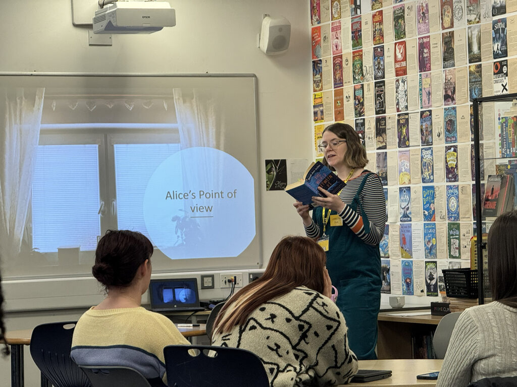 A speaker stands at the front of a classroom reading aloud from a book, with a projector screen behind them showing the text "Alice's Point of view".