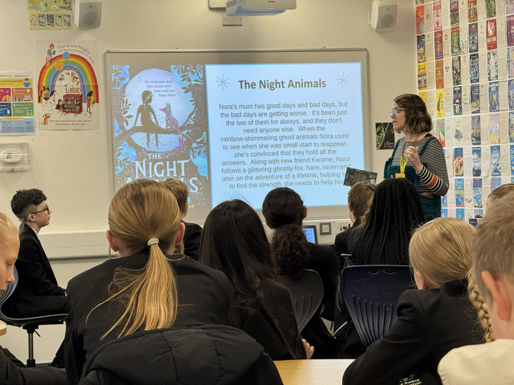 A speaker holds a copy of the book The Night Animals while presenting a slide about the story to a group of students in a classroom.