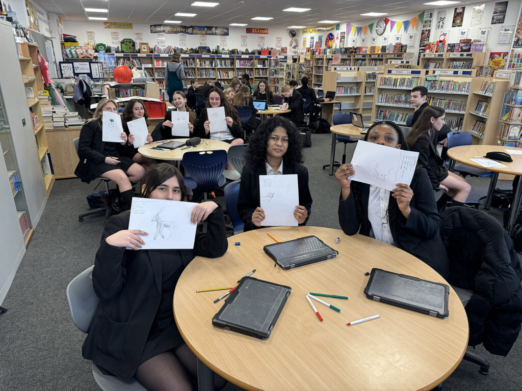 A group of students sitting around a table in a library, proudly holding up pencil drawings of animals they have created.