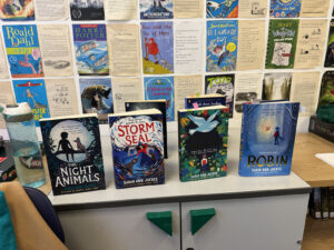 Four children's books by author Sarah Ann Juckes are displayed on a counter, with a large collage of classic book covers decorating the wall behind them.
